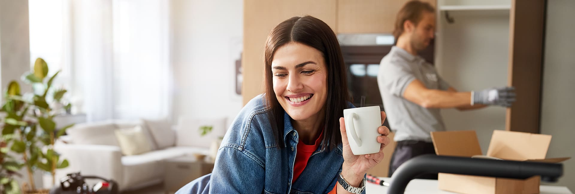Frau hält eine Tasse, während im Hintergrund ein Monteur Möbel aufbaut.